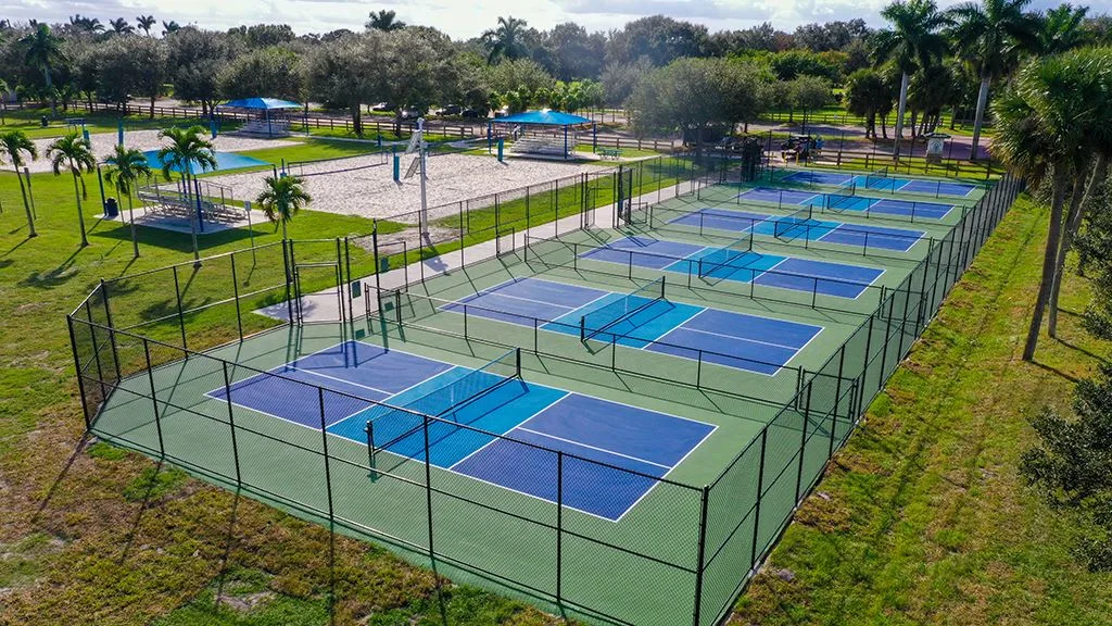 Aerial view of six blue and green pickleball courts at Greenbriar Park in Wellington, with sand volleyball courts and palm trees behind them